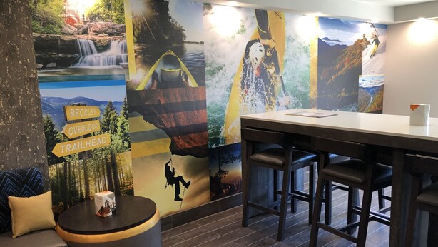 A modern bar area with a wooden bar counter and stools, a round table with a coffee cup, and a mural with outdoor scenes and directional signs.
