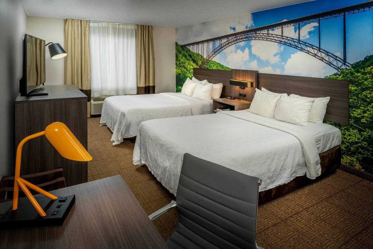 A hotel room with two beds, a desk with a lamp, a TV, and a mural of a bridge.