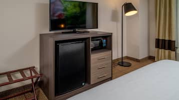 Hotellihuone, jossa on seinälle asennettu taulutelevisio, joka on kiinnitetty puiseen kaappiin, pieni jääkaappi, lamppu ja valkoisella peitteellä varustettu sänky.