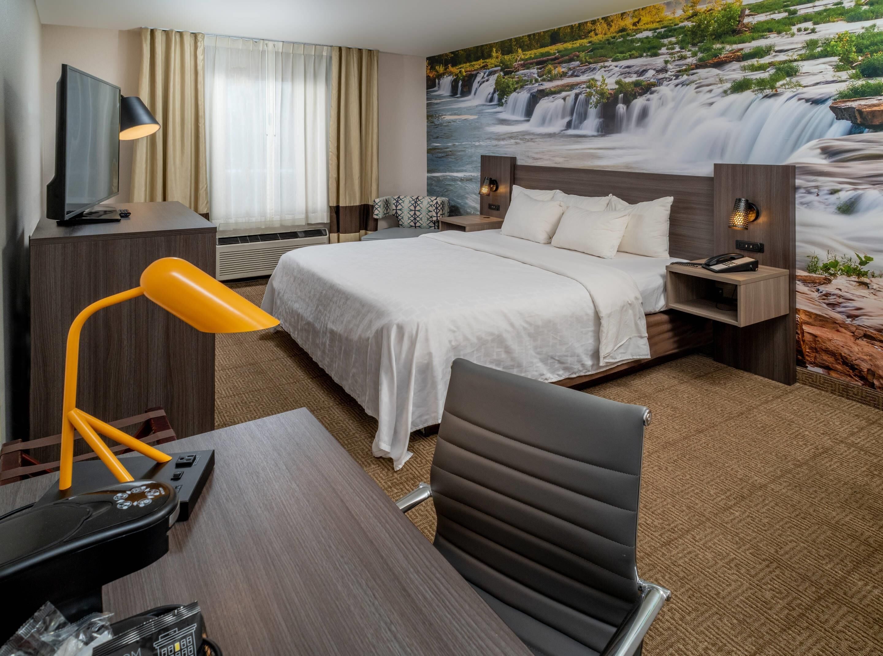 A hotel room with a bed, a desk, a chair, a TV, and a mural of a waterfall.