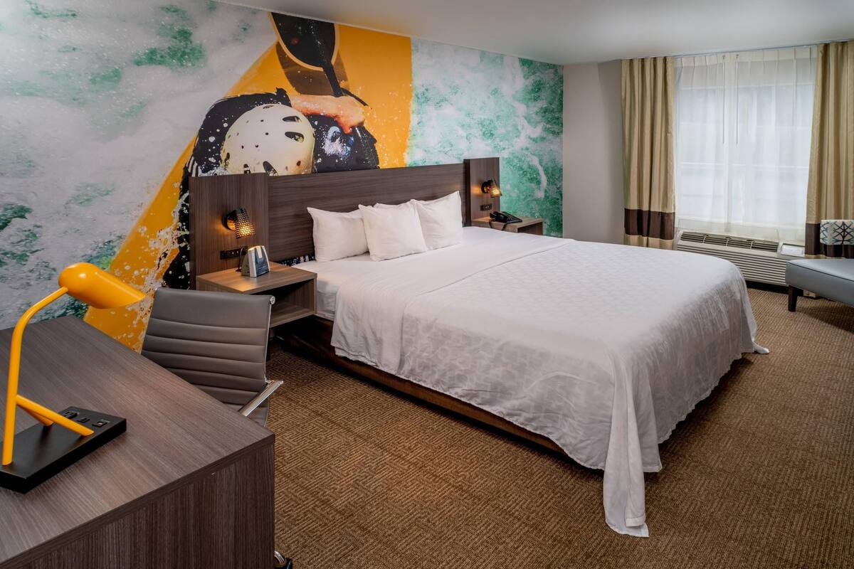 A hotel room with a large bed, a desk with a lamp, a nightstand, and a wall with a colorful mural.