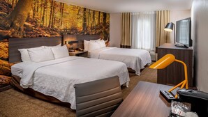 A hotel room with two beds, a desk, a chair, a TV, and a wall mural of a forest.
