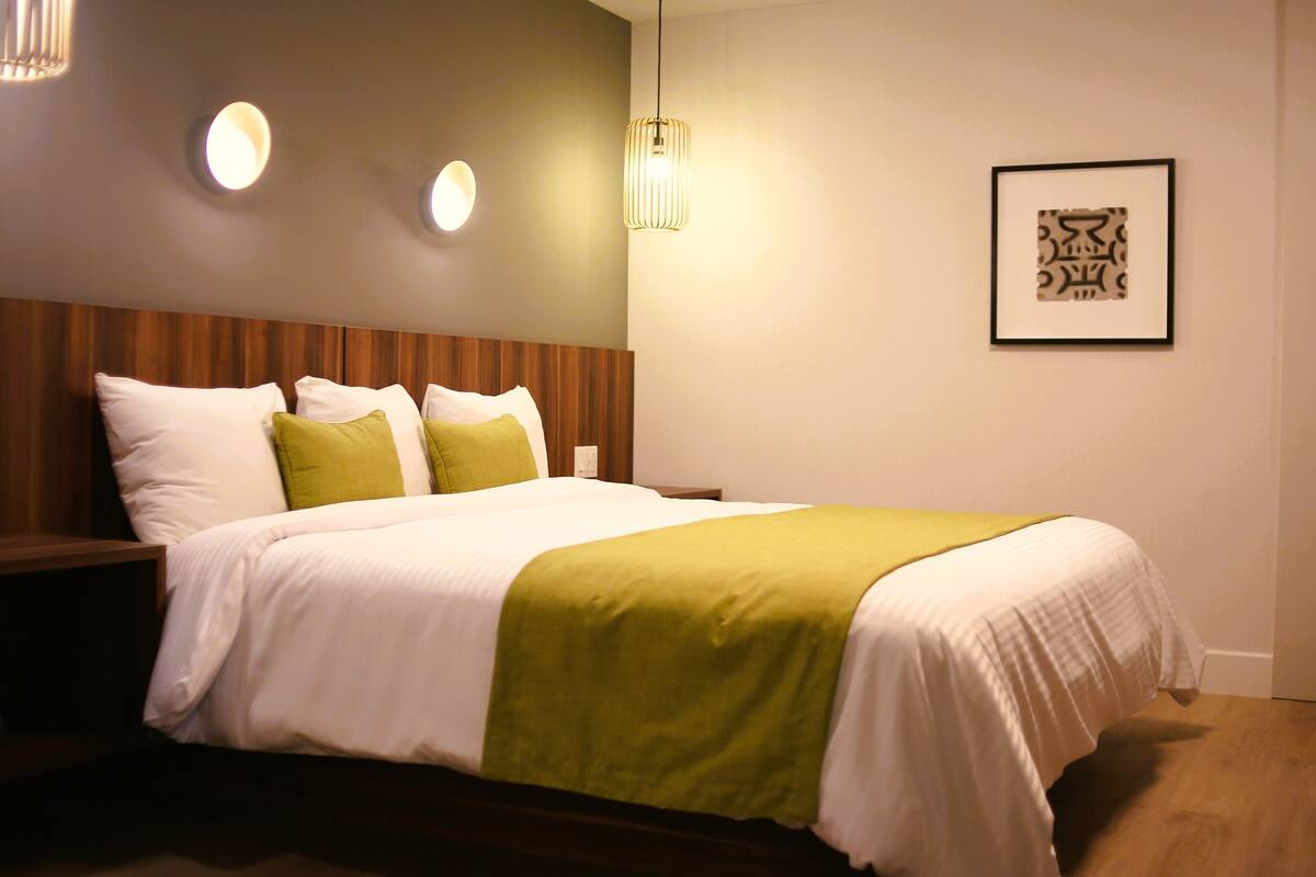 A neatly made bed with a green throw, a wooden headboard, and two wall-mounted lights.