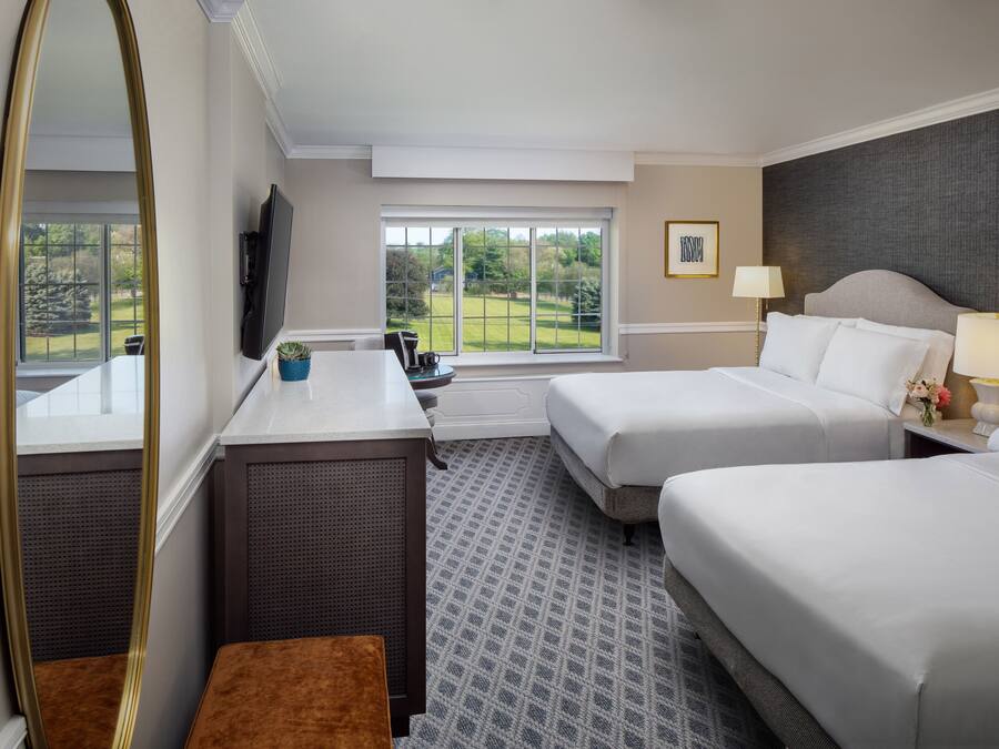 A hotel room with two beds, a desk, a chair, a large mirror, a TV, and a window with a view of greenery.