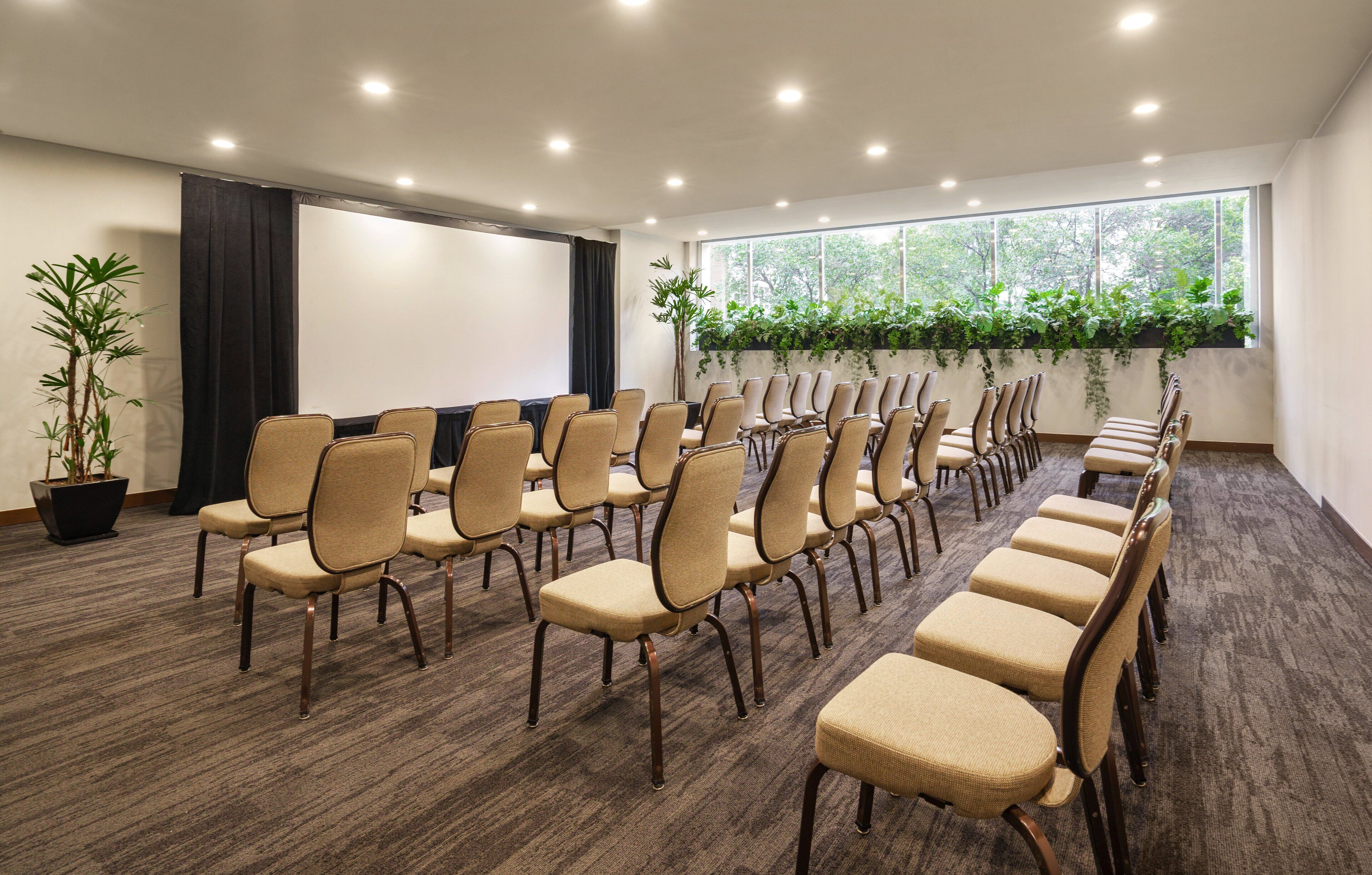 A conference room with rows of chairs, a projector screen, and a large window with plants.