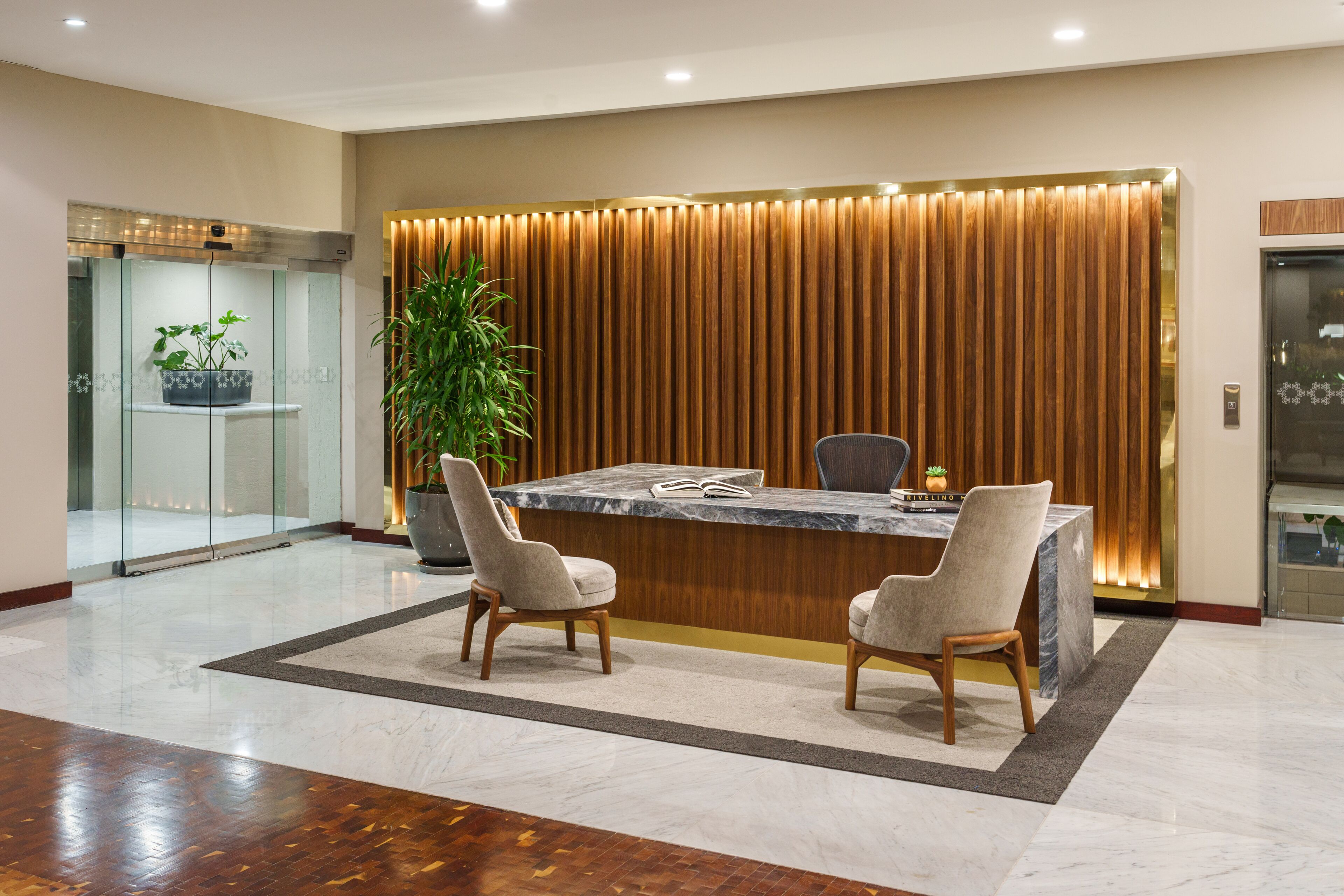 A reception area with a marble reception desk, two upholstered chairs, a large potted plant, and a wooden panel wall with vertical slats.