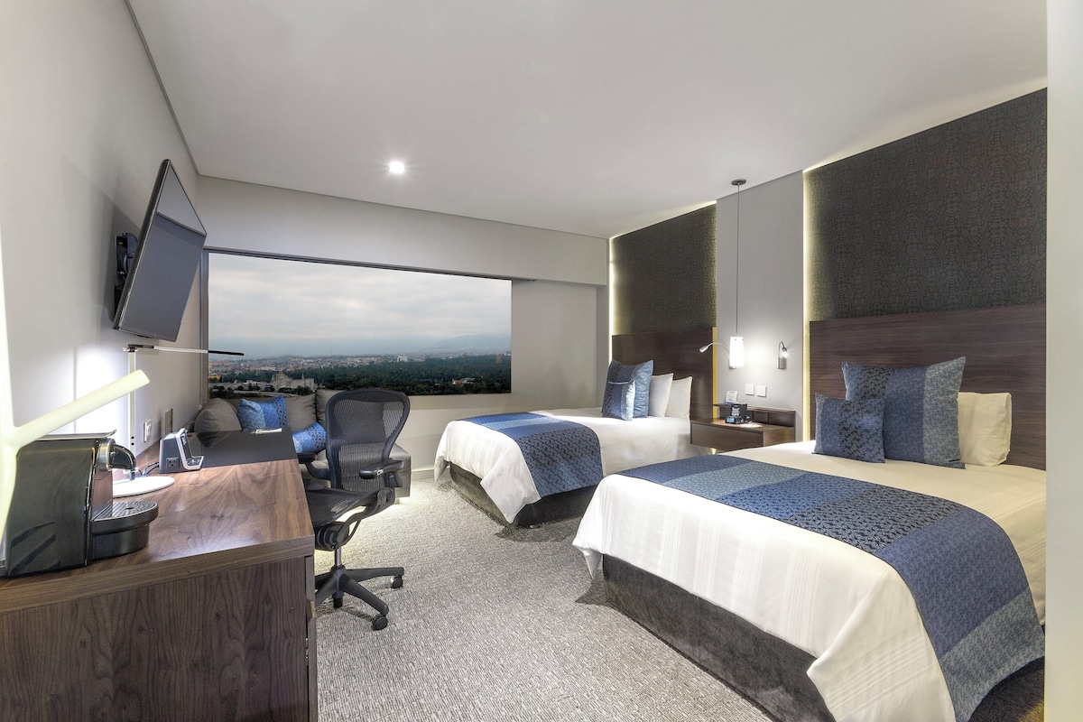 Habitación de hotel con dos camas, un escritorio con computadora, una silla, un televisor montado en la pared y un ventanal con vista a la ciudad.