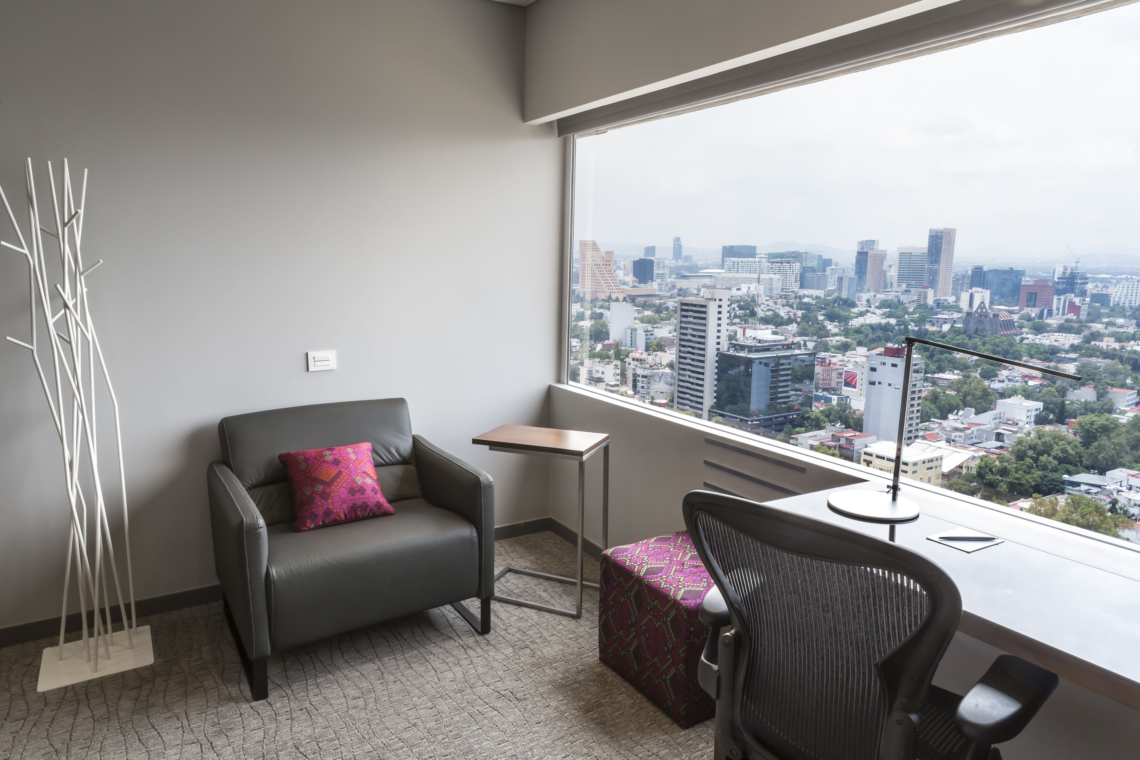 A modern hotel room with a city view, a gray sofa with a pink pillow, a small table, and a black office chair.