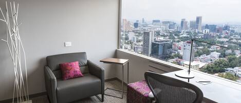 A modern hotel room with a city view, a gray sofa with a pink pillow, a small table, and a black office chair.