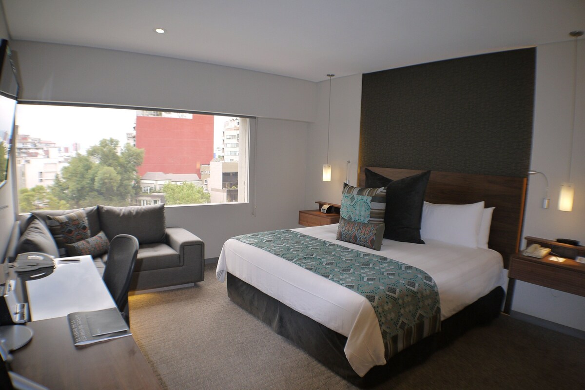 Habitación de hotel con una cama grande, un sofá, un escritorio y una ventana con vista a edificios.
