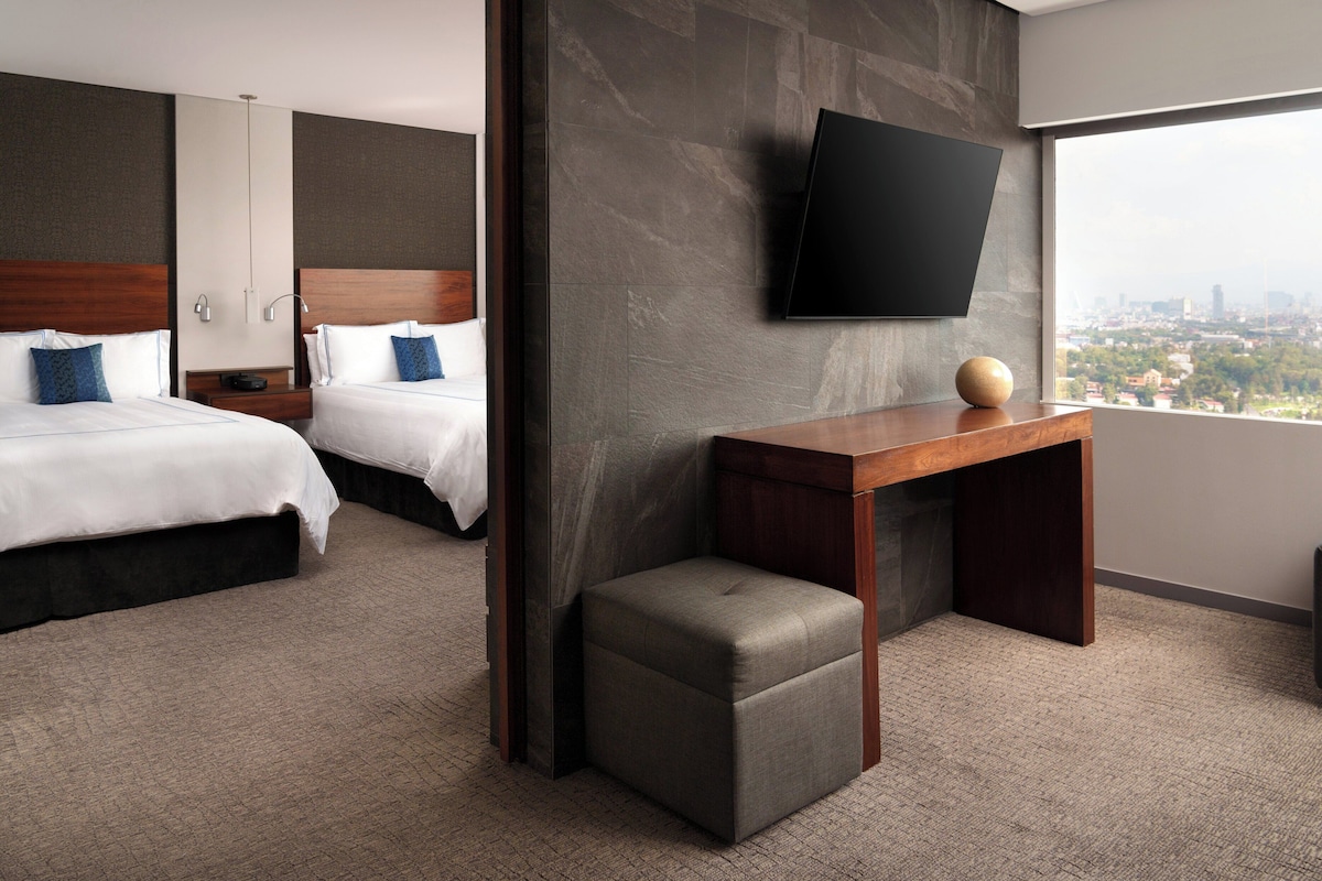 Habitación de hotel con dos camas, un escritorio, una silla, un televisor montado en la pared y un ventanal con vistas a la ciudad.