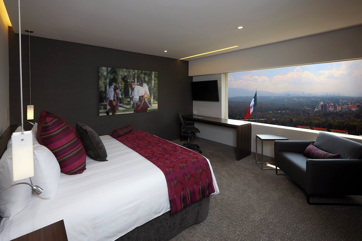 Habitación de hotel moderna con una cama grande, un escritorio, una silla y vistas a un paisaje urbano.