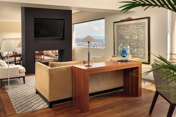 A modern living room with a sofa, a coffee table, a TV mounted on the wall, and a large window offering a city view.