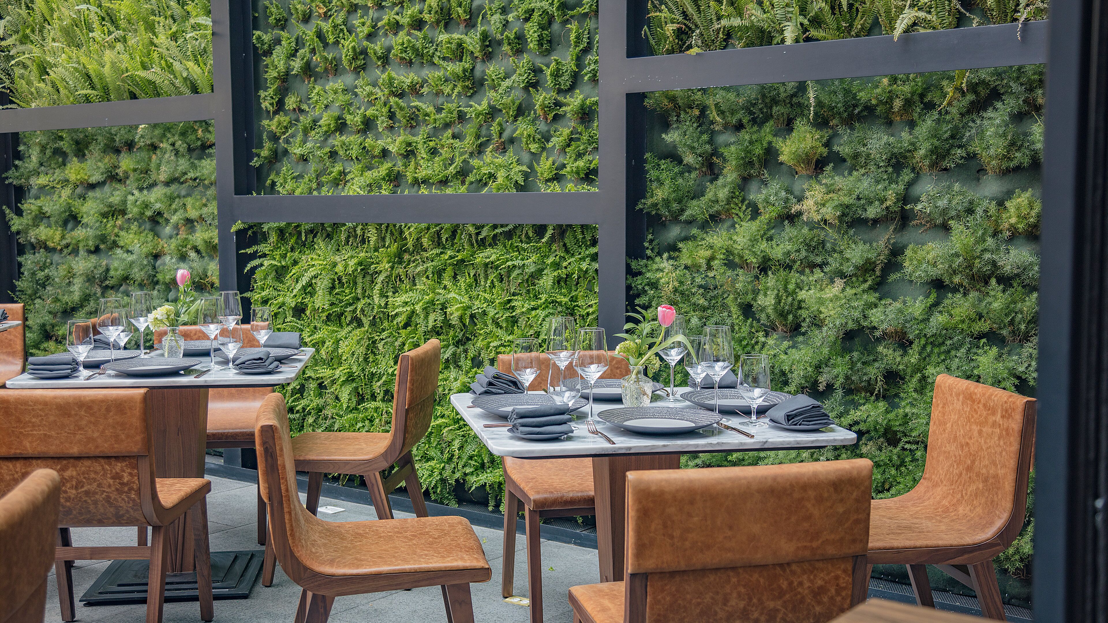 Un área de comedor al aire libre con mesas dispuestas para dos, sillas y una pared verde exuberante al fondo.