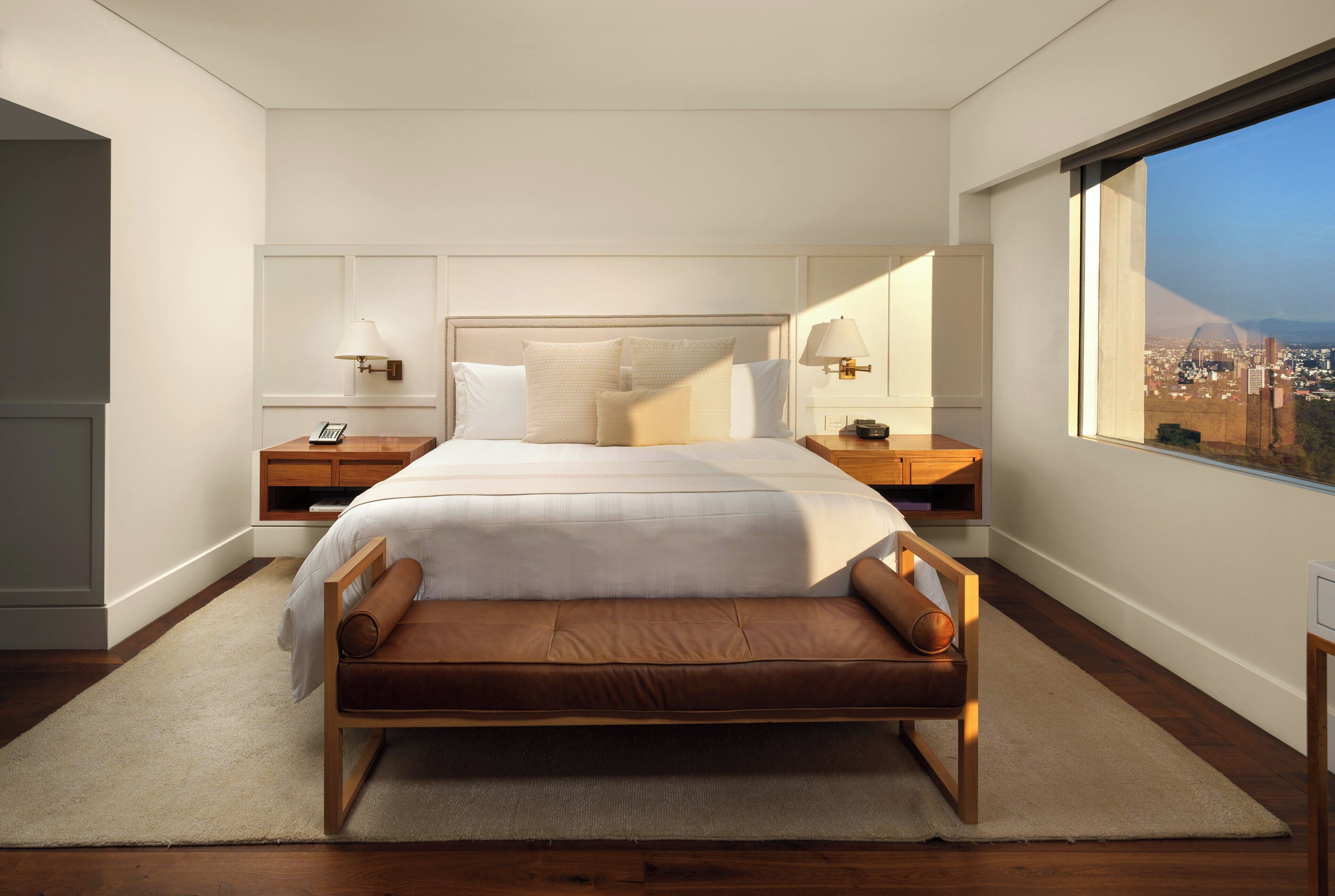 A modern hotel room with a large bed, wooden headboard, two bedside tables with lamps, a view of a cityscape through a large window, and a city skyline visible outside.