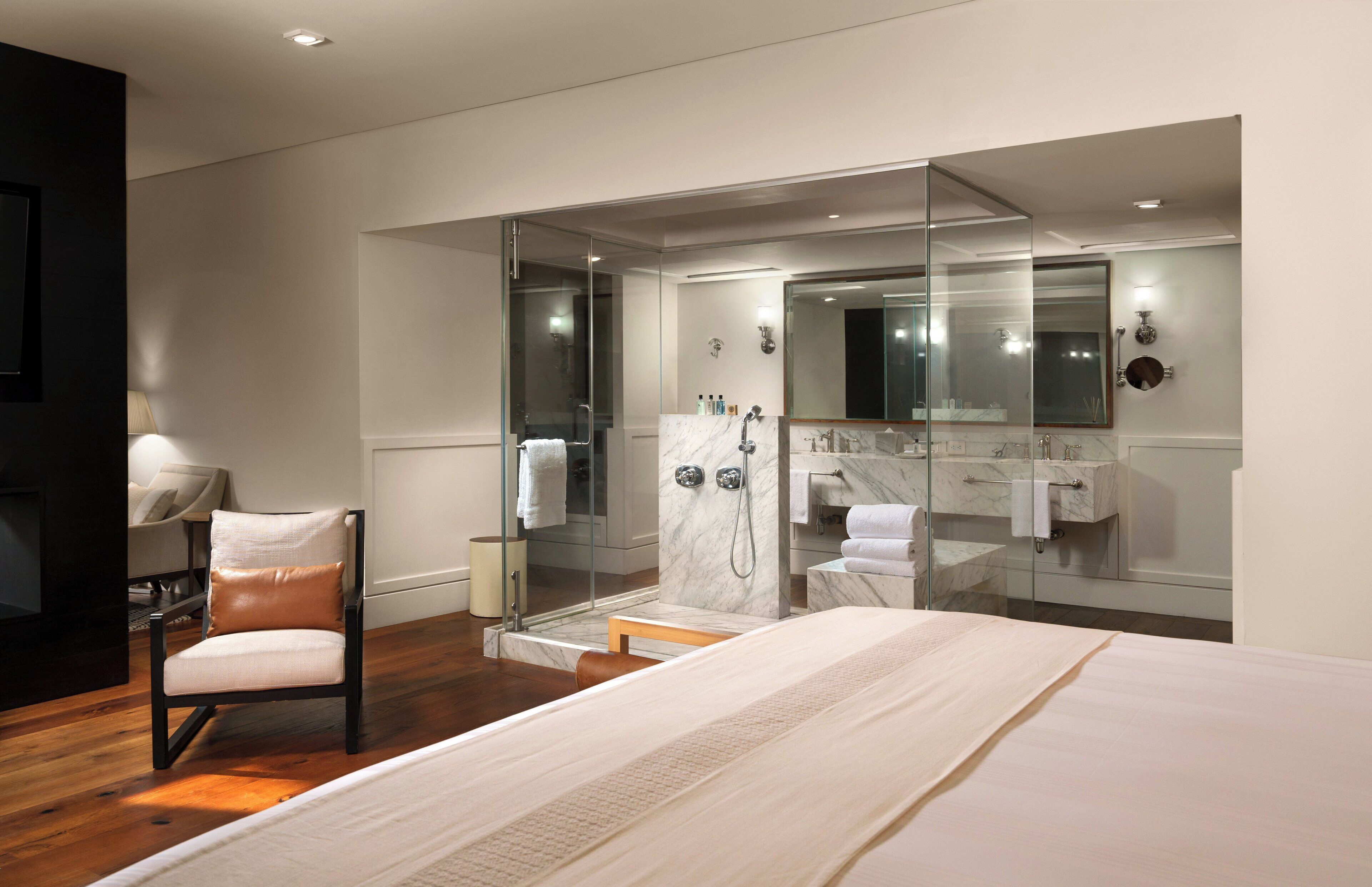 A modern hotel room with a large bed, a glass-enclosed shower, a vanity with a mirror, and a seating area with a chair.