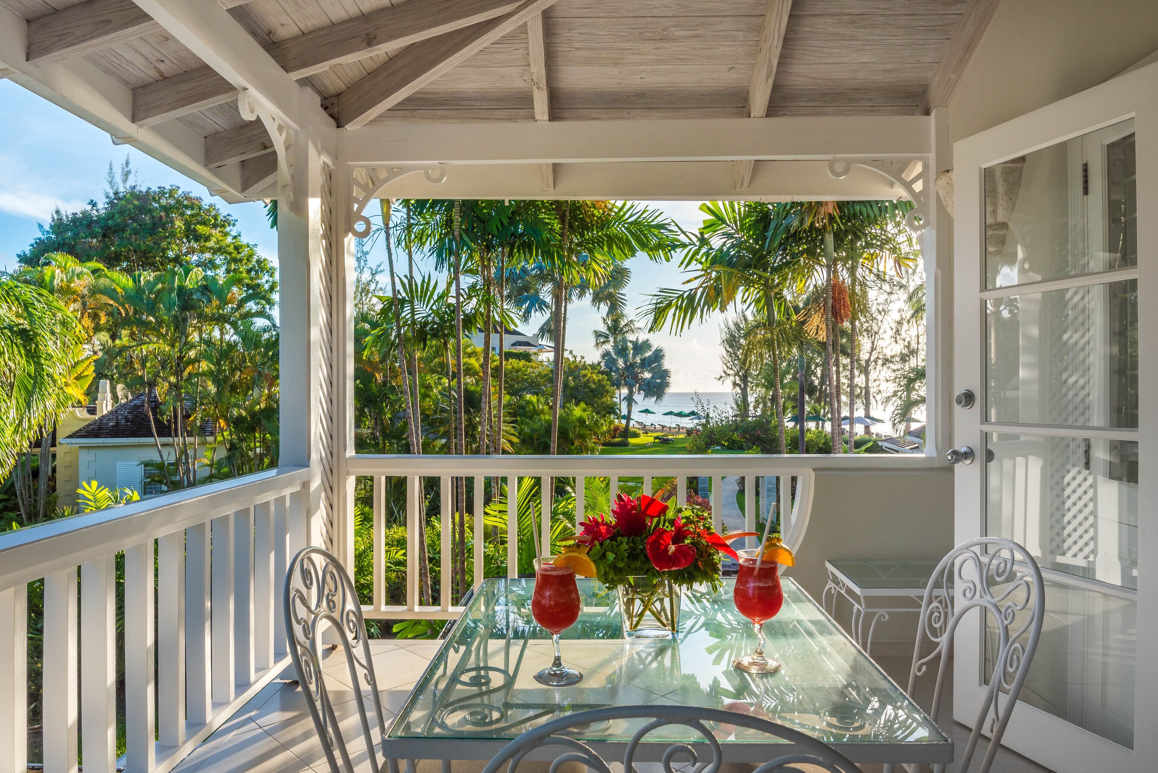 A glass table with two drinks and a flower arrangement on a balcony with a white railing, overlooking a tropical garden and a body of water.