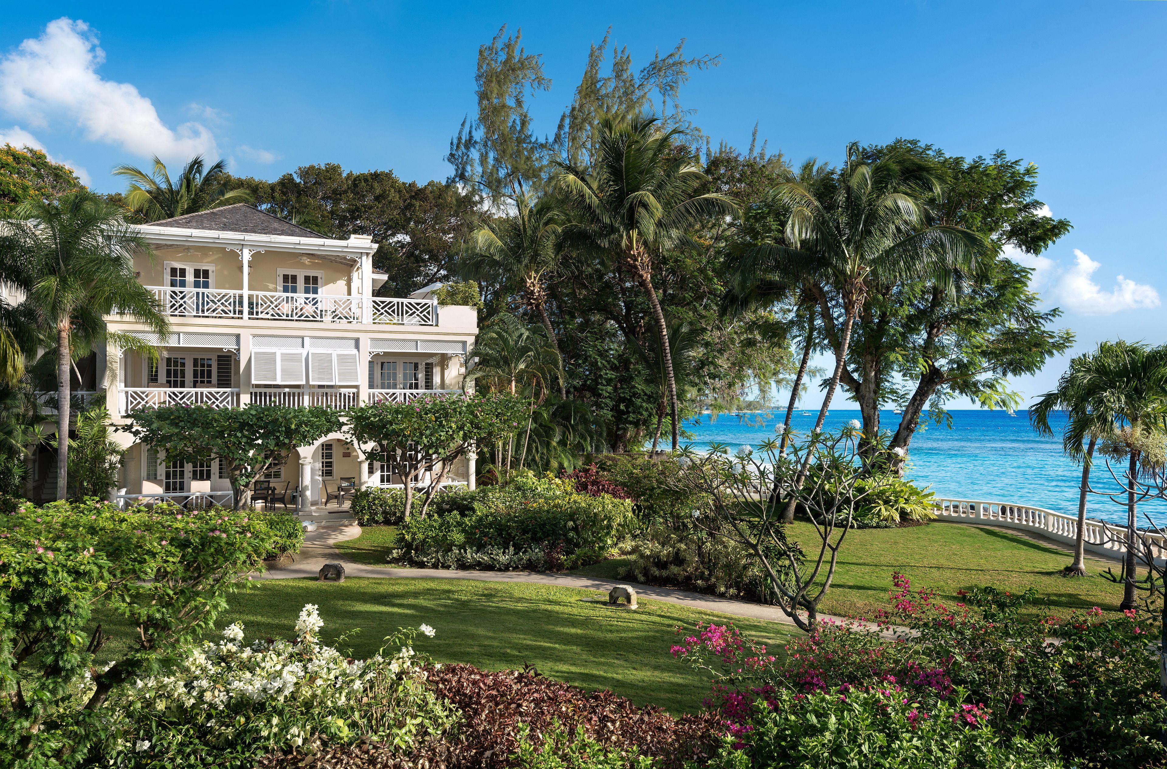 A large, two-story house with a balcony, surrounded by lush greenery and a view of the ocean.