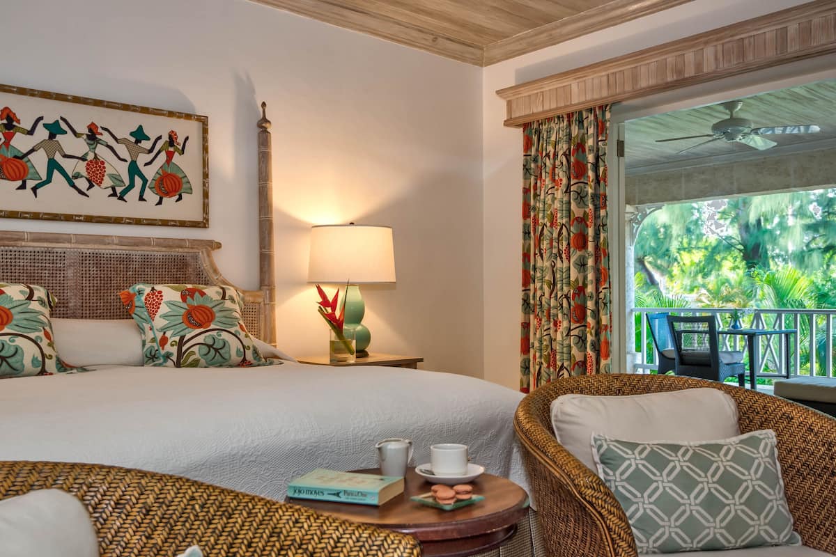 A hotel room with a bed, a nightstand with a lamp, a wicker chair, a small table with a book and a cup, and a view of the outdoors.