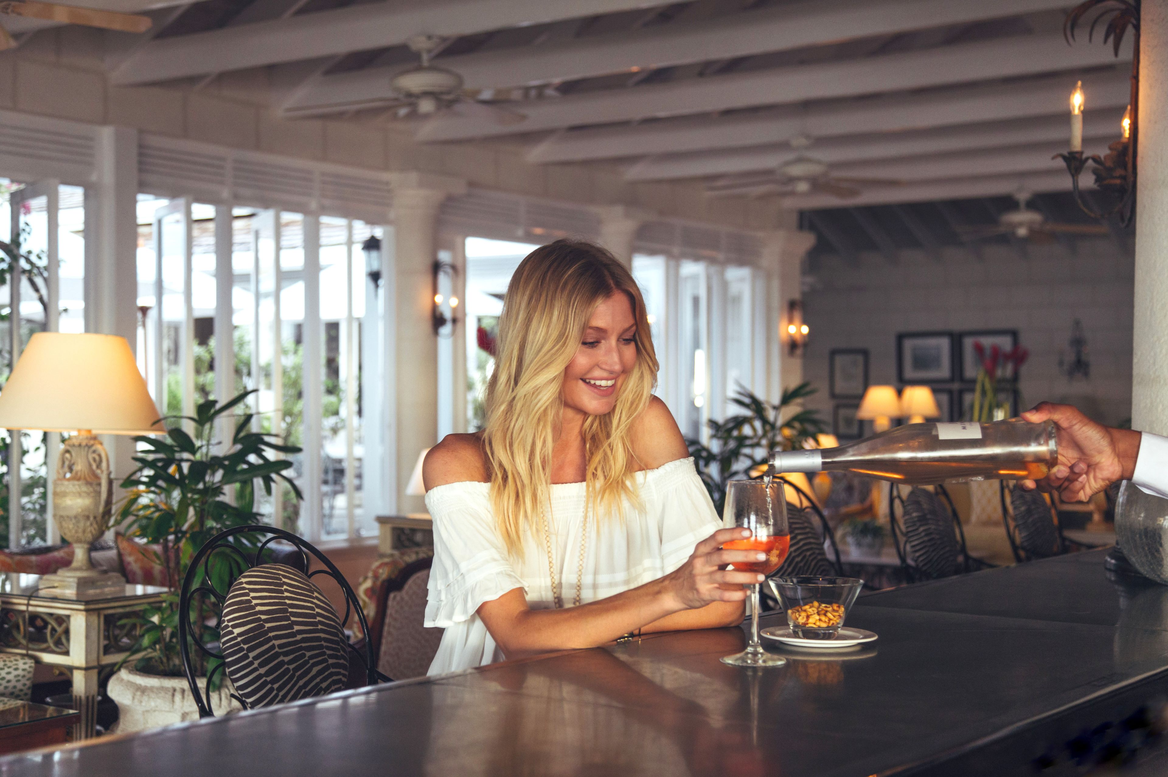 A woman seated at a bar in a well-lit room with large windows, a chandelier, and a plant in the background.