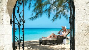 A beachside lounge chair with a view of the ocean, a decorative metal gate, and a palm tree. - Coral Reef Club (Holetown)
