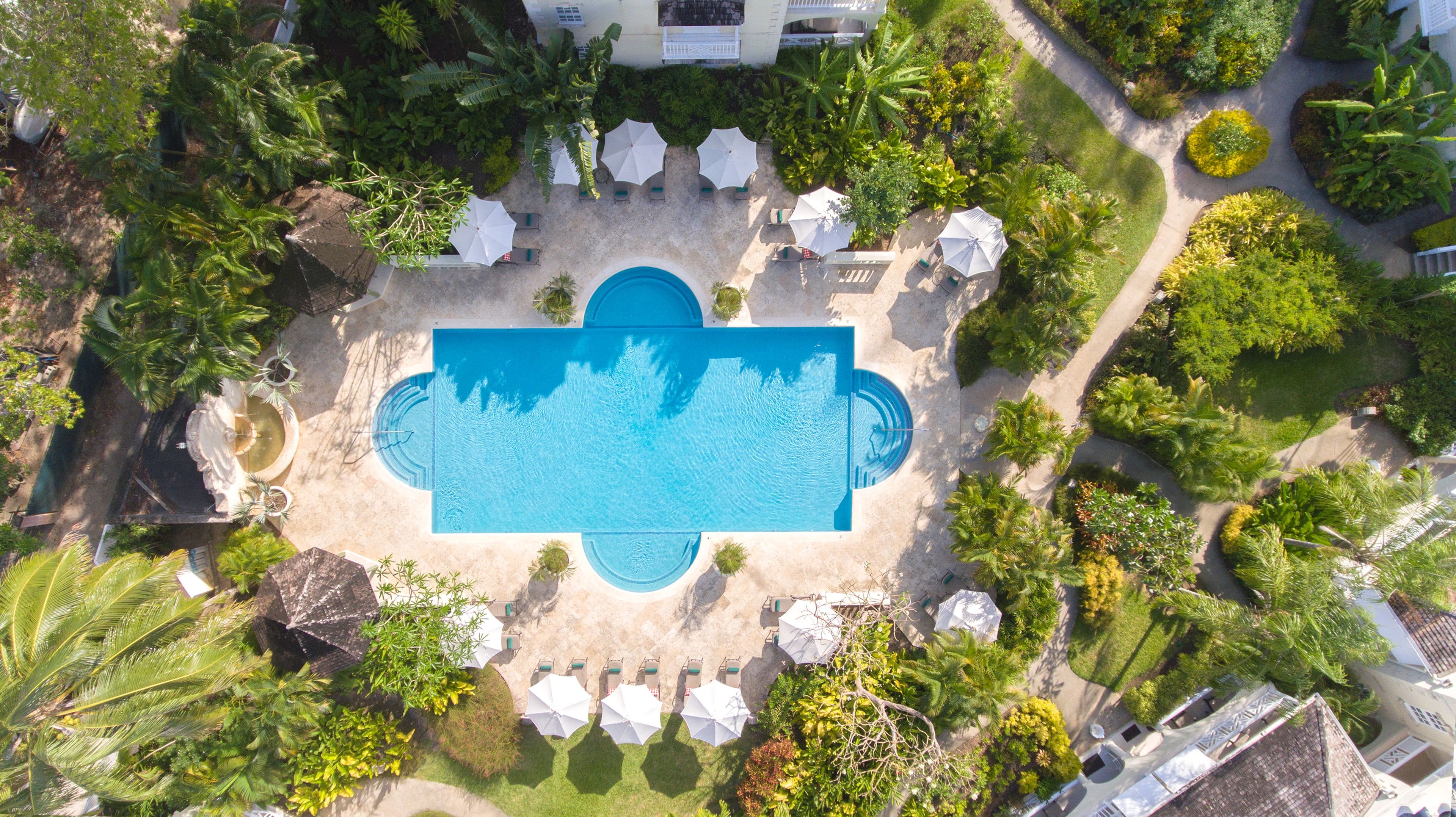 Vista aérea de un complejo turístico con una piscina central, rodeada de sillas de descanso y sombrillas, y abundante vegetación.