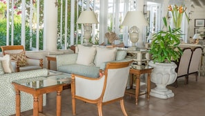 A well-lit, spacious room with a large window, a sofa set, a glass coffee table, and a potted plant.