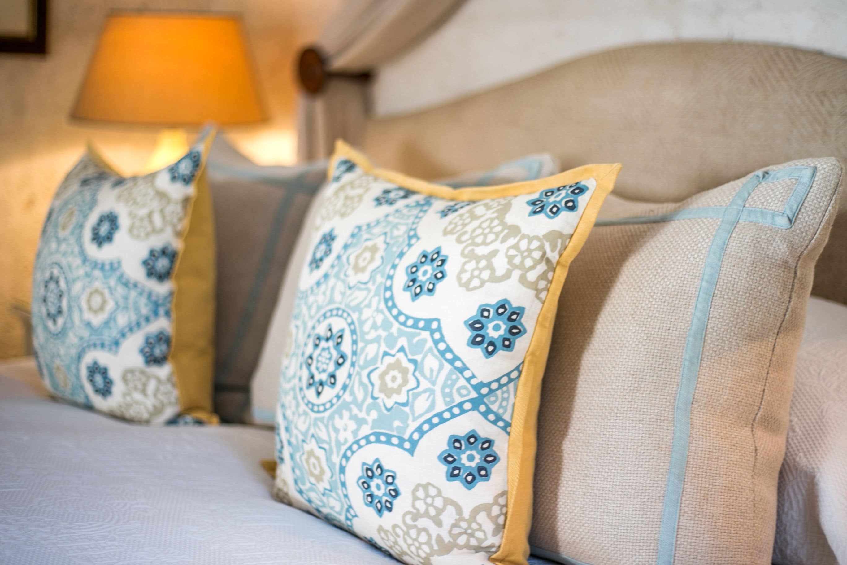 Una cama con dos almohadas de estampado floral azul y blanco, cabecera color beige y una lámpara al fondo.