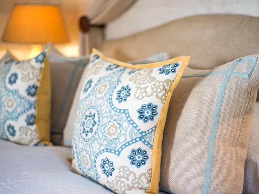 Una cama con dos almohadas de estampado floral azul y blanco, cabecera color beige y una lámpara al fondo.
