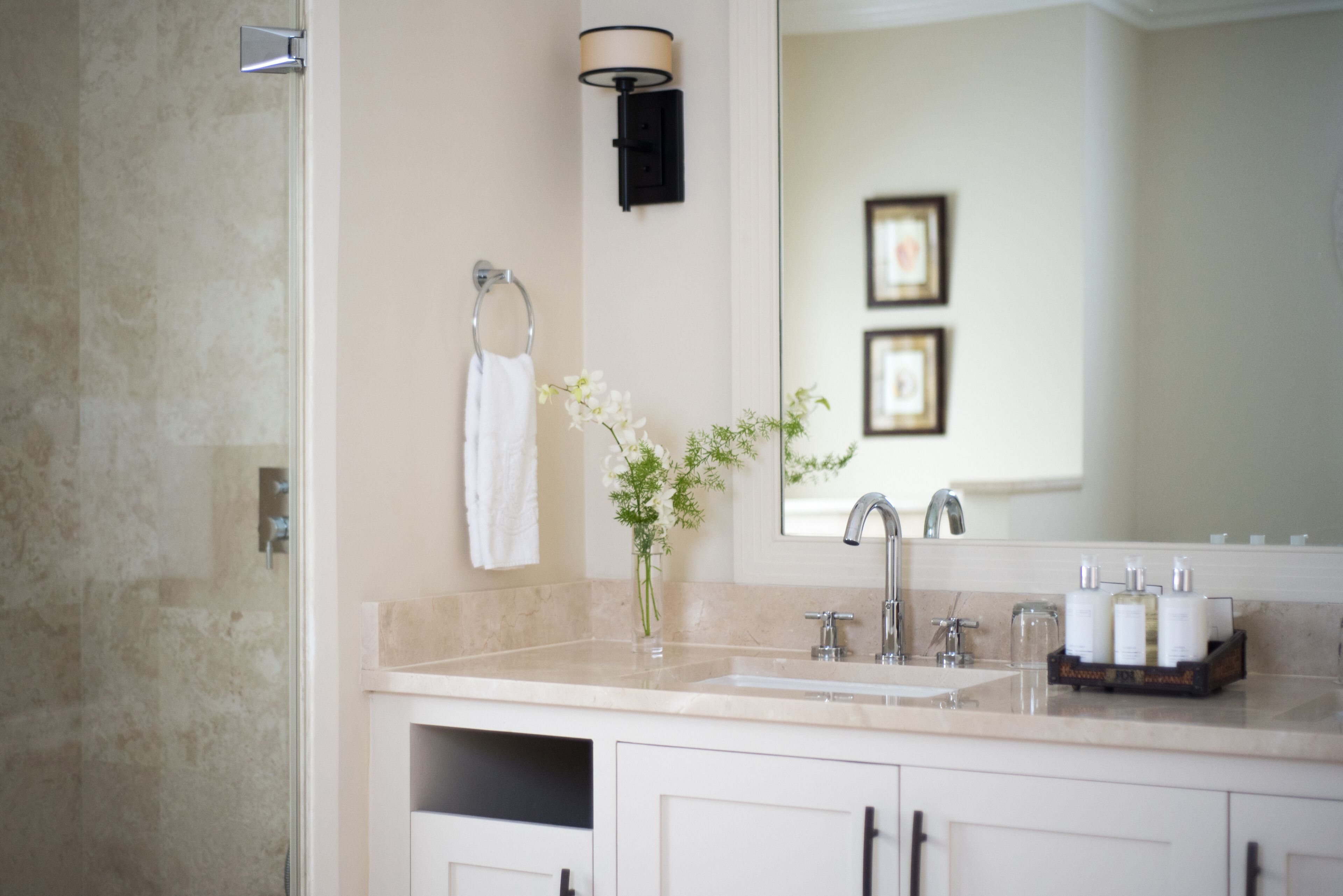 A modern bathroom with a beige countertop, a white sink, a mirror, and a vase with flowers.