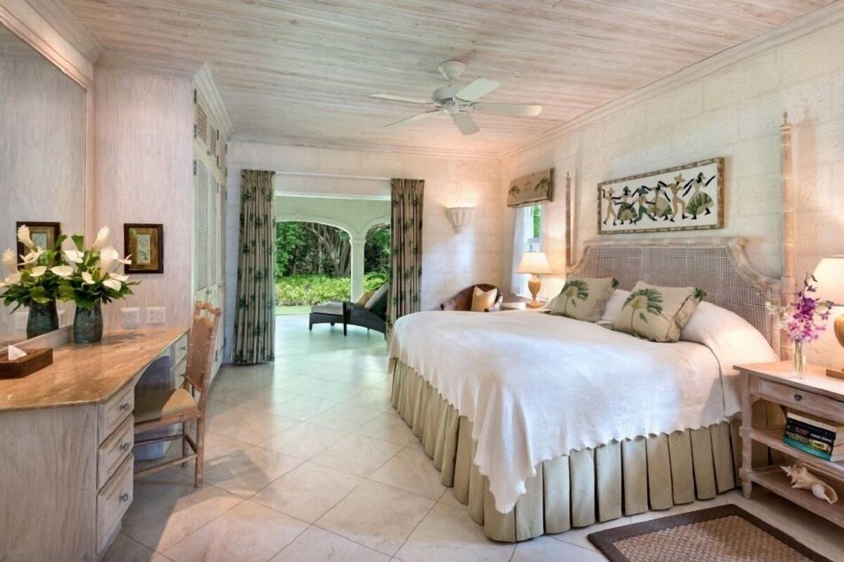 A bedroom with a large bed, a desk, and a chair. There is a window with curtains, a ceiling fan, and a painting on the wall.