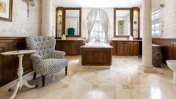 A spacious bathroom with a large central sink, two vanity mirrors, a patterned armchair, and a decorative vase.