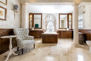 A spacious bathroom with a large central sink, two vanity mirrors, a patterned armchair, and a decorative vase.