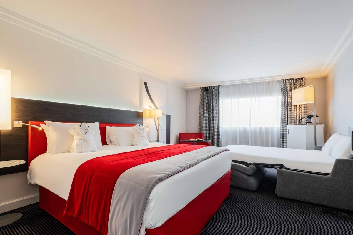 A hotel room with a large bed, a smaller bed, a desk, a chair, and a window with curtains.