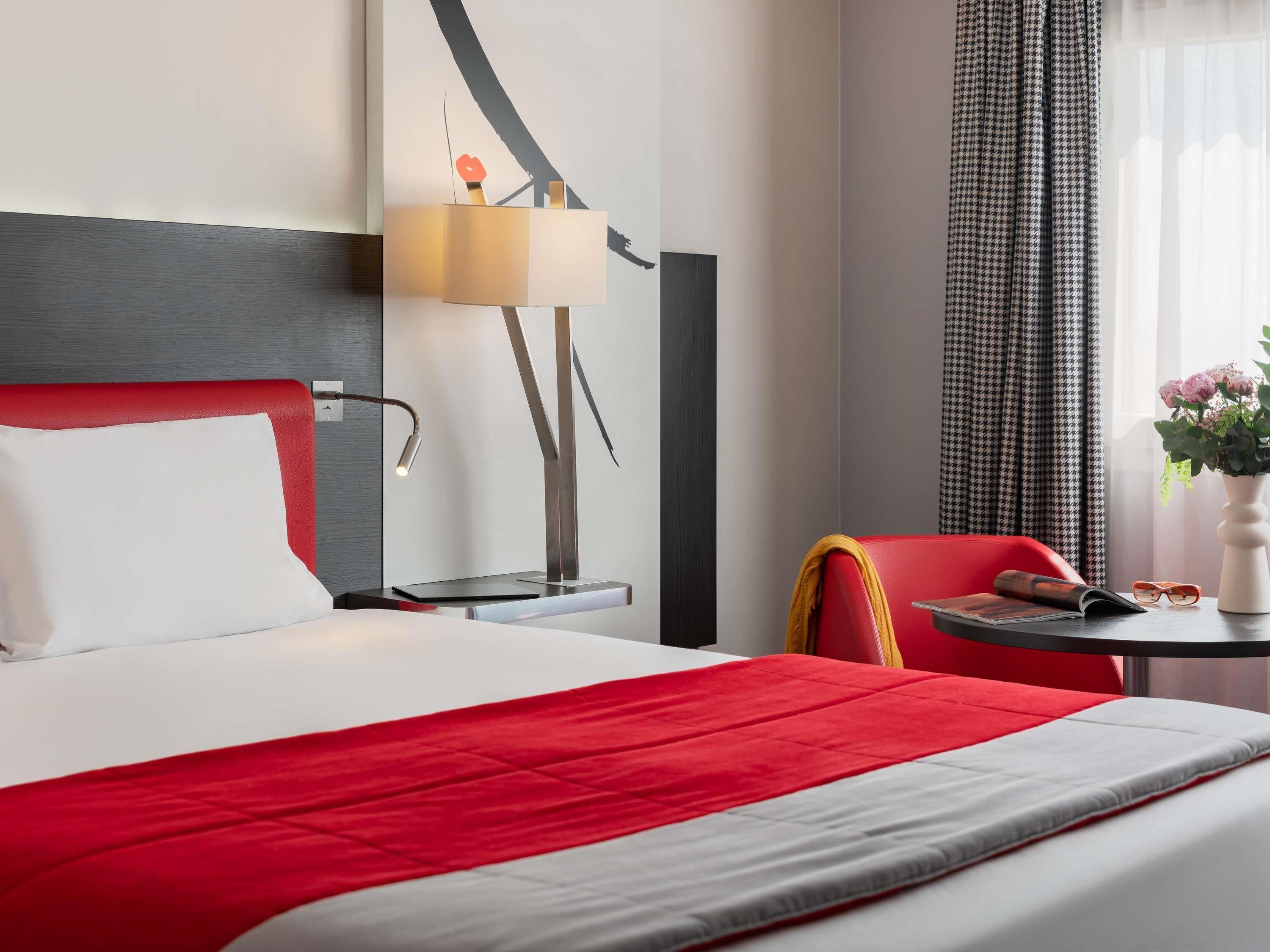 Ein Hotelzimmer mit einem Bett, einem roten Sessel, einem kleinen Tisch mit Blumenvase und einer Lampe.