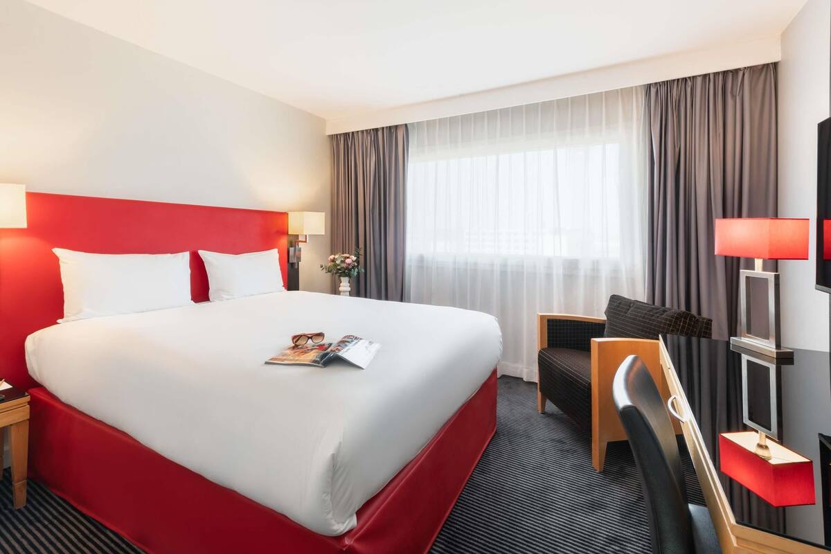 A hotel room with a large bed, a desk, a chair, a TV, and a window with curtains.