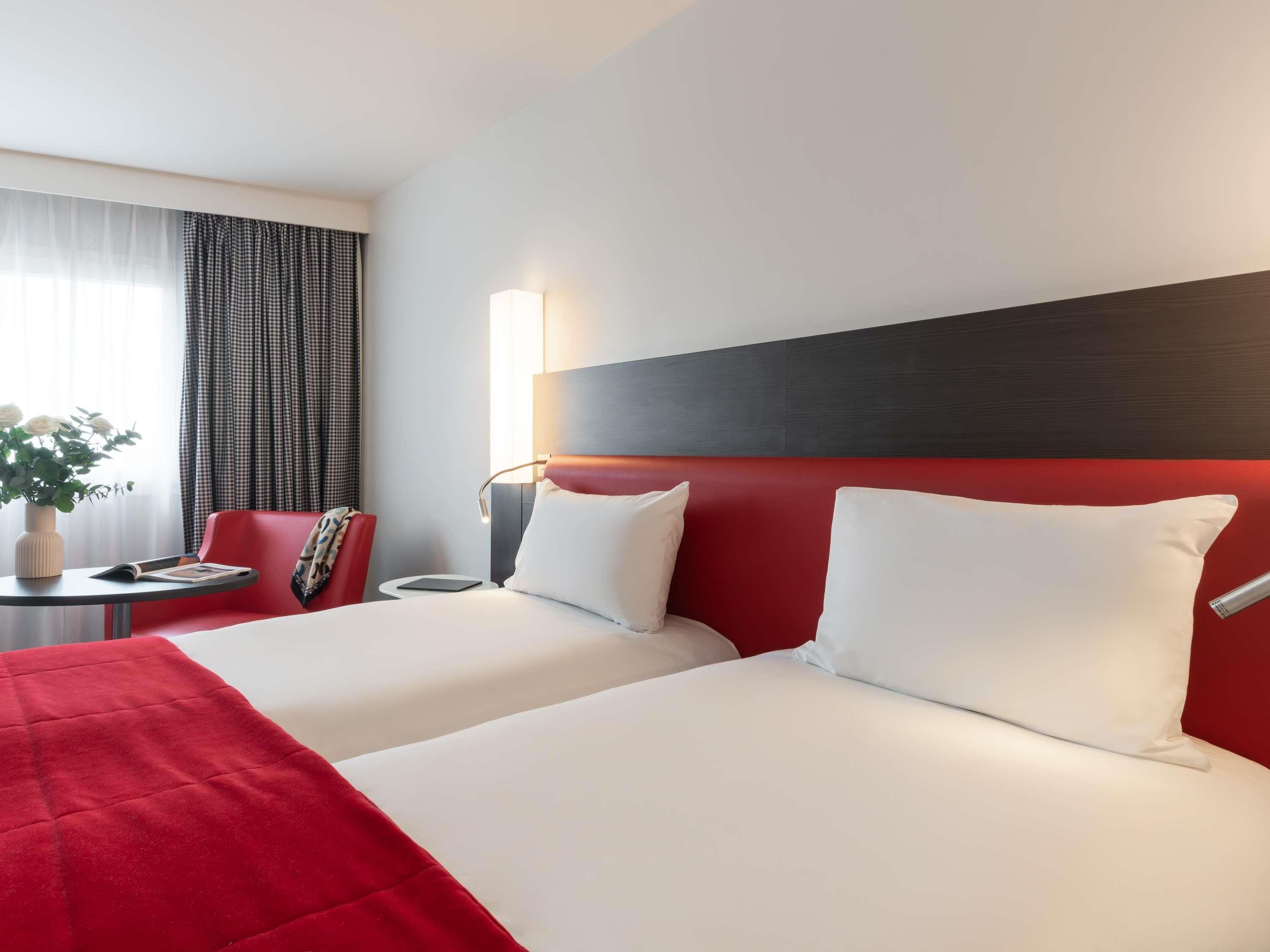 A hotel room with two beds, a red headboard, a bedside table with a lamp, a small table with a vase of flowers, and a chair.
