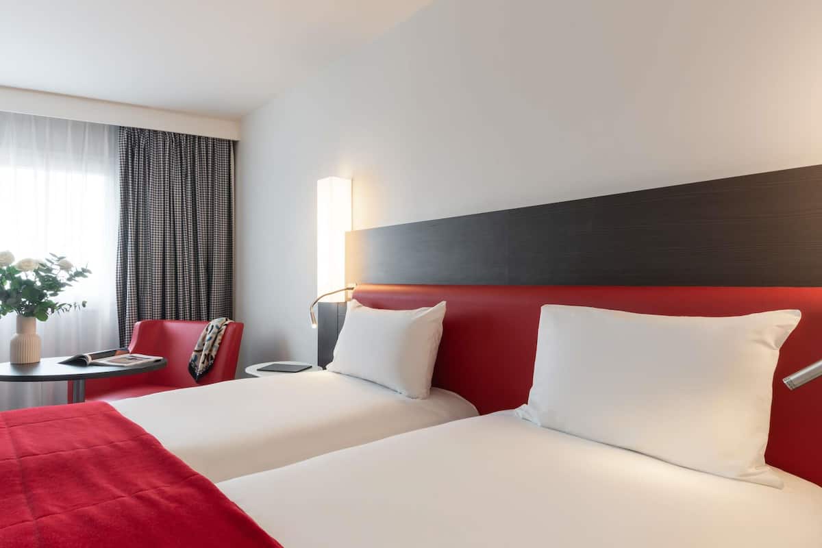 A hotel room with two beds, a red headboard, a bedside table with a lamp, a small table with a vase of flowers, and a chair.