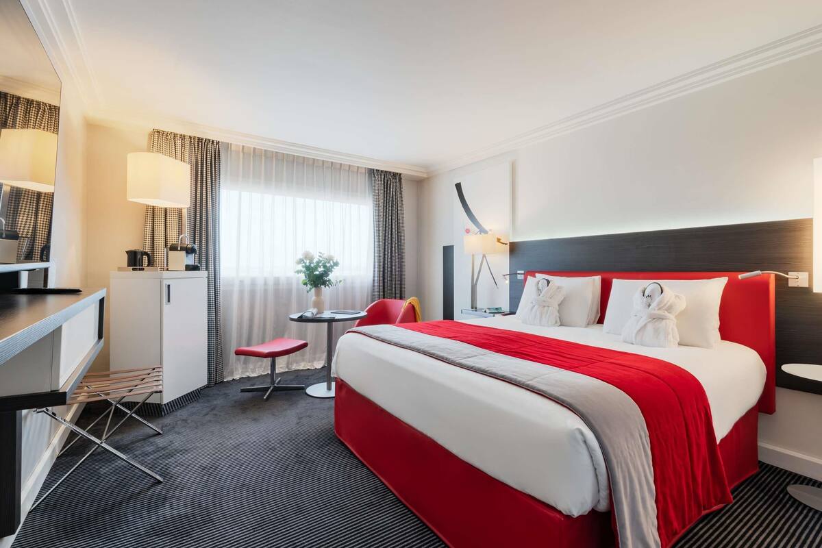 A hotel room with a bed, a desk, a chair, a small table with a vase of flowers, and a window with curtains.