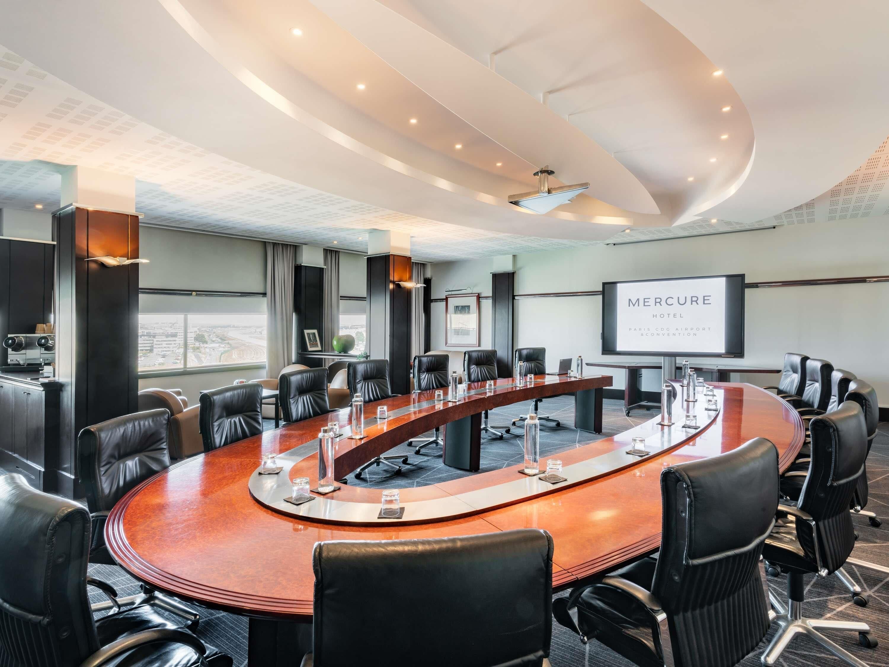 Un salón de conferencias con una larga mesa, sillas de cuero negro, una pantalla de proyector y vistas a la ciudad.