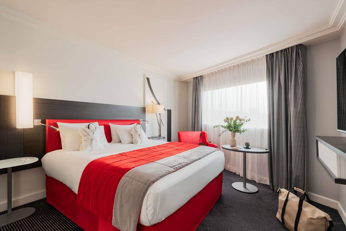 A hotel room with a large bed, a bedside table, a chair, a small table with a vase of flowers, and a mirror on the wall.