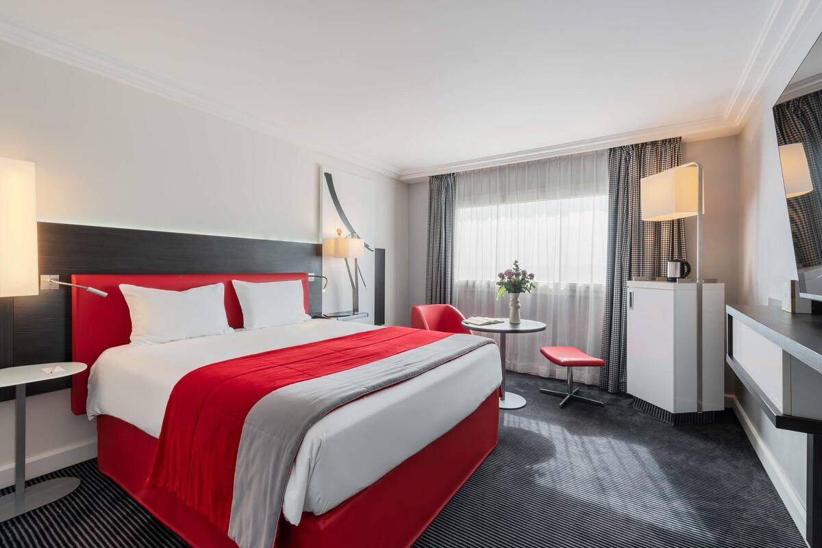 A hotel room with a large bed, a desk, a chair, a small table with a vase, and a TV.