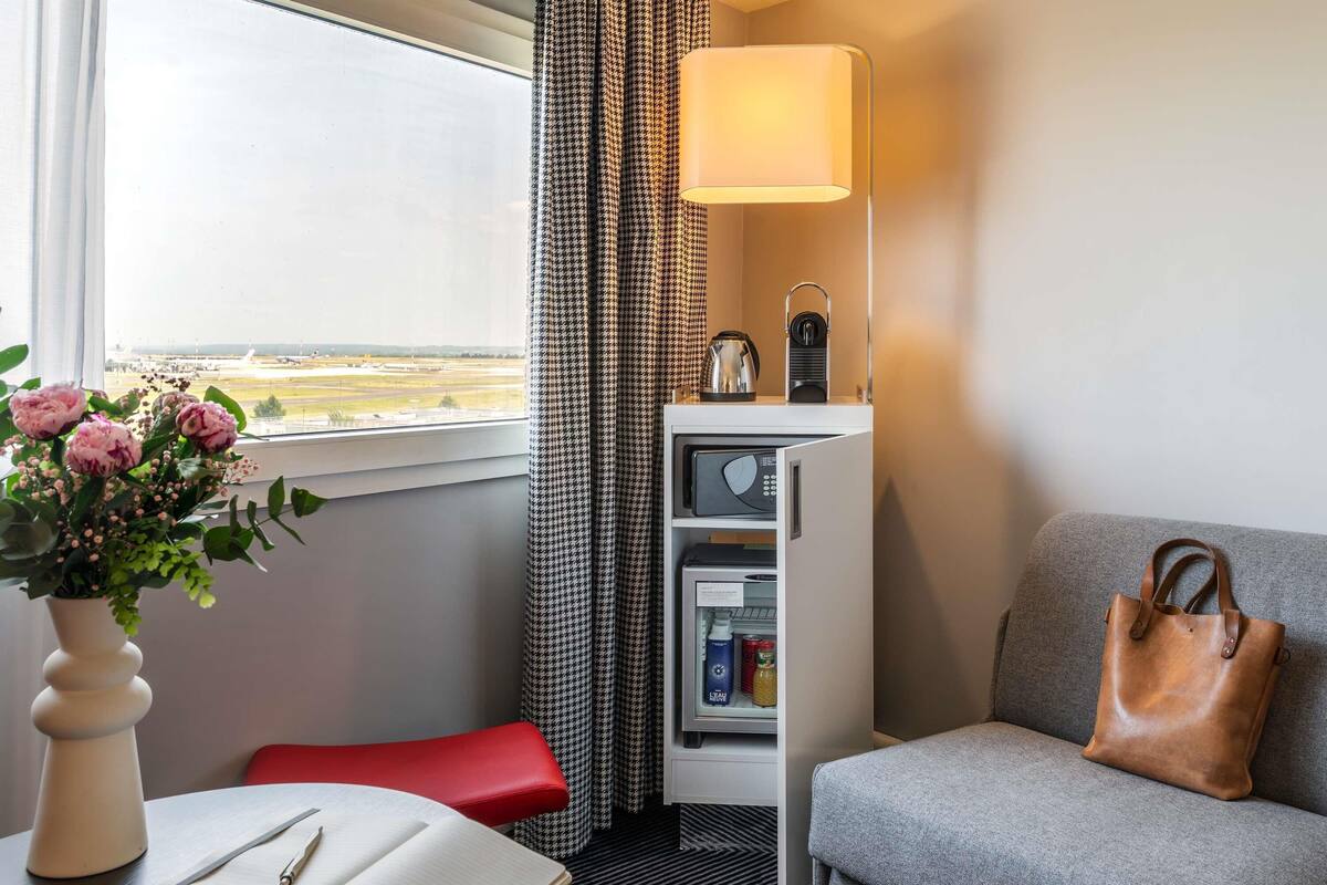 A hotel room with a sofa, a table with a book and a vase of flowers, a microwave, a refrigerator, and a view of an airport.