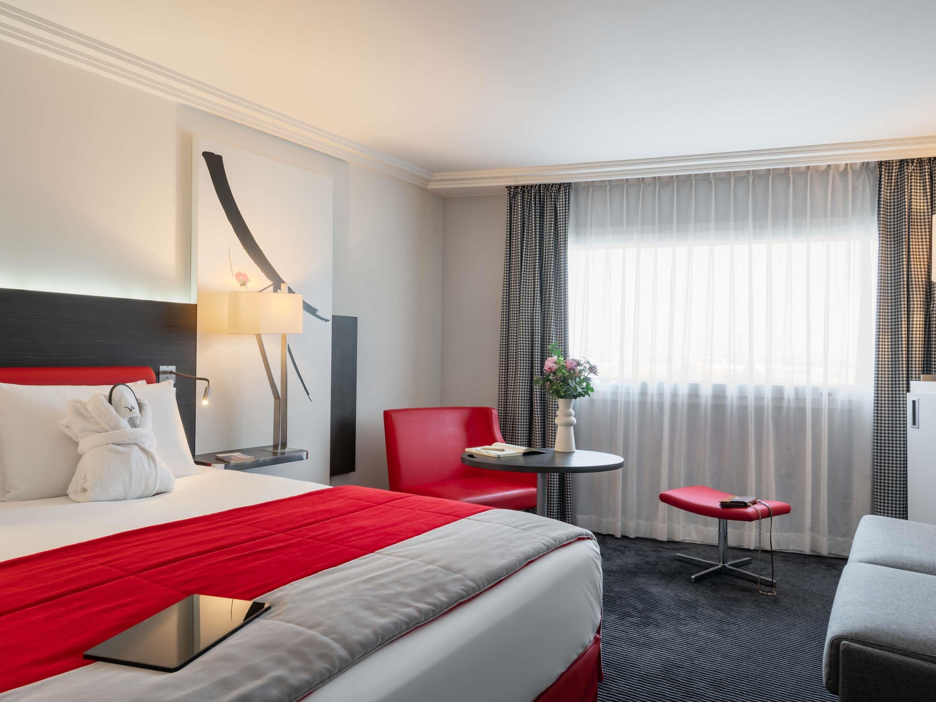 Une chambre d’hôtel avec un lit, un bureau, une chaise, une petite table et une fenêtre avec des rideaux.