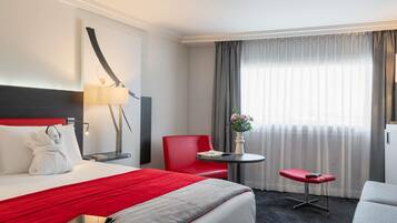 Une chambre d’hôtel avec un lit, un bureau, une chaise, une petite table et une fenêtre avec des rideaux.