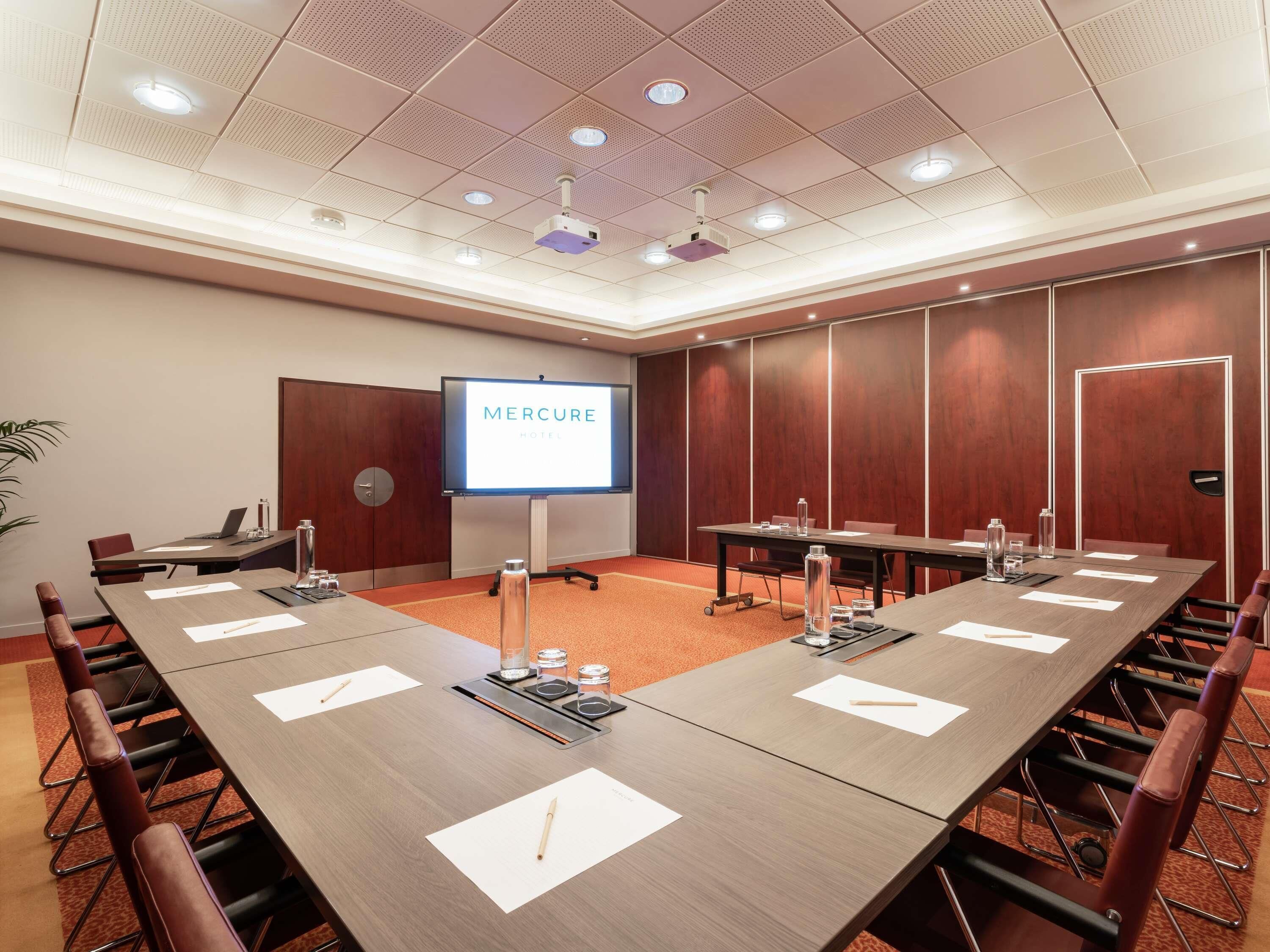 Un salón de conferencias con una larga mesa, sillas, una pantalla de proyector que muestra 'MERCURE HOTEL' y una pared de paneles de madera.