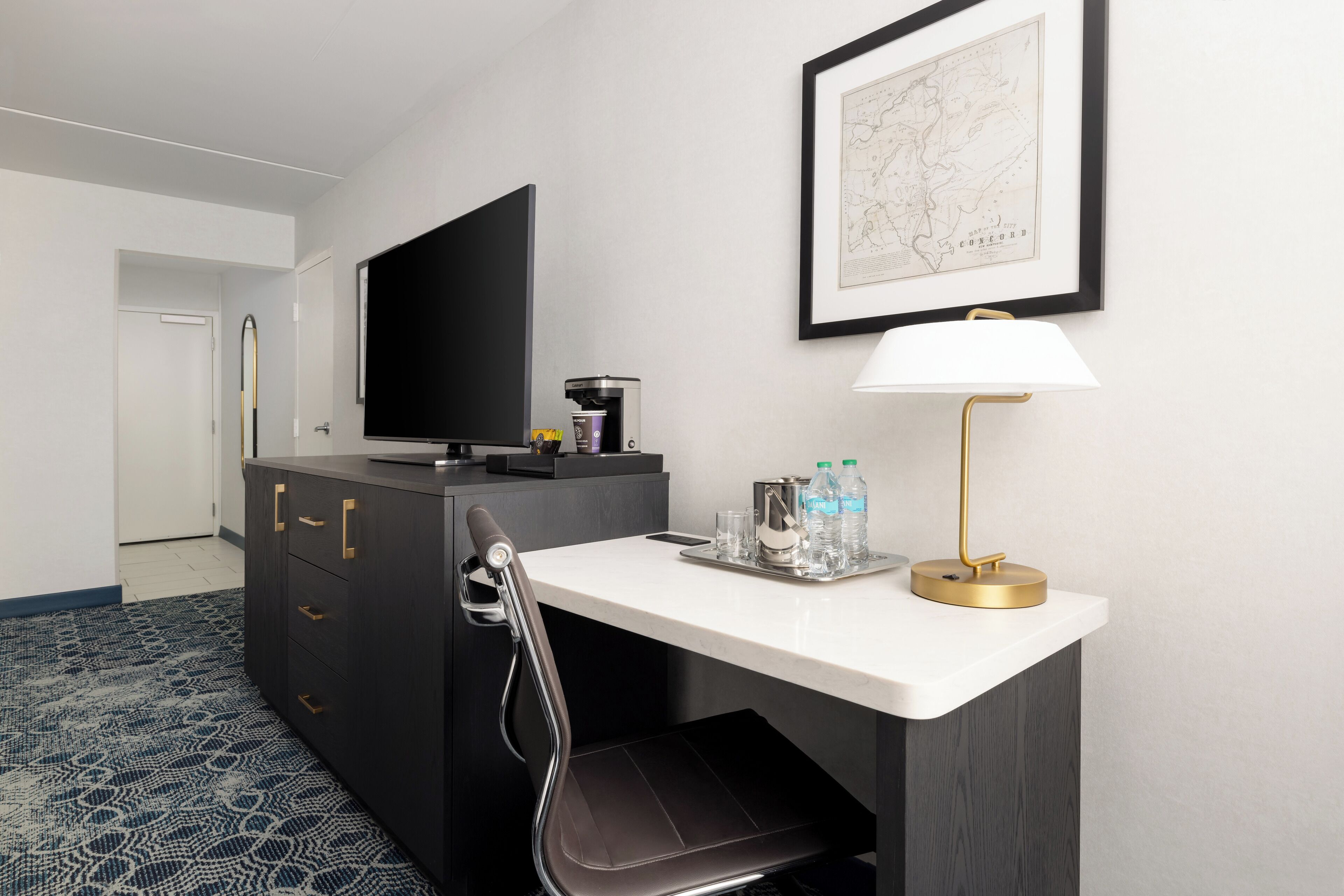 A hotel room with a desk, a chair, a television, a map on the wall, and a coffee machine.