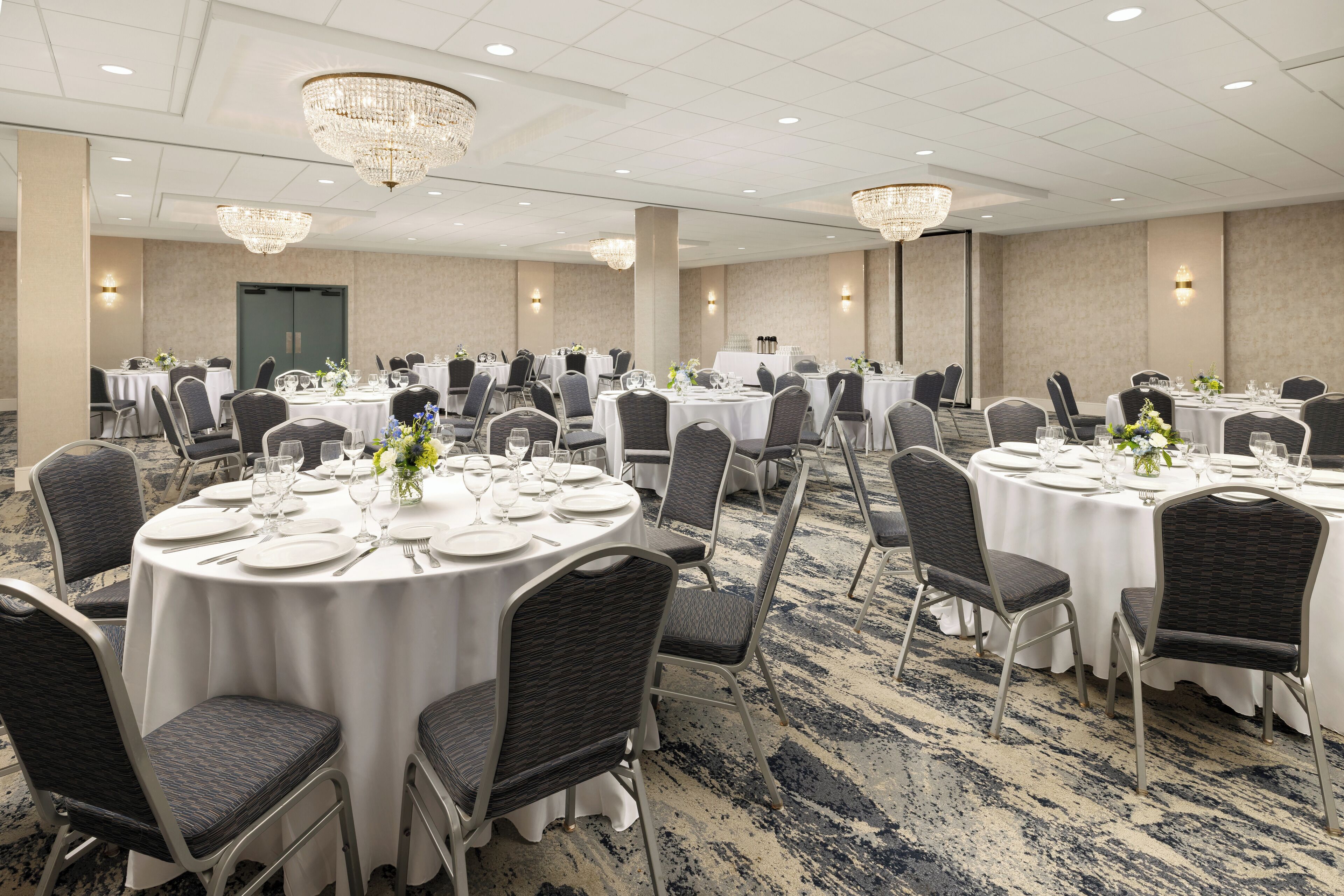 A banquet hall with round tables set for dining, featuring white tablecloths, dark chairs, and chandeliers.