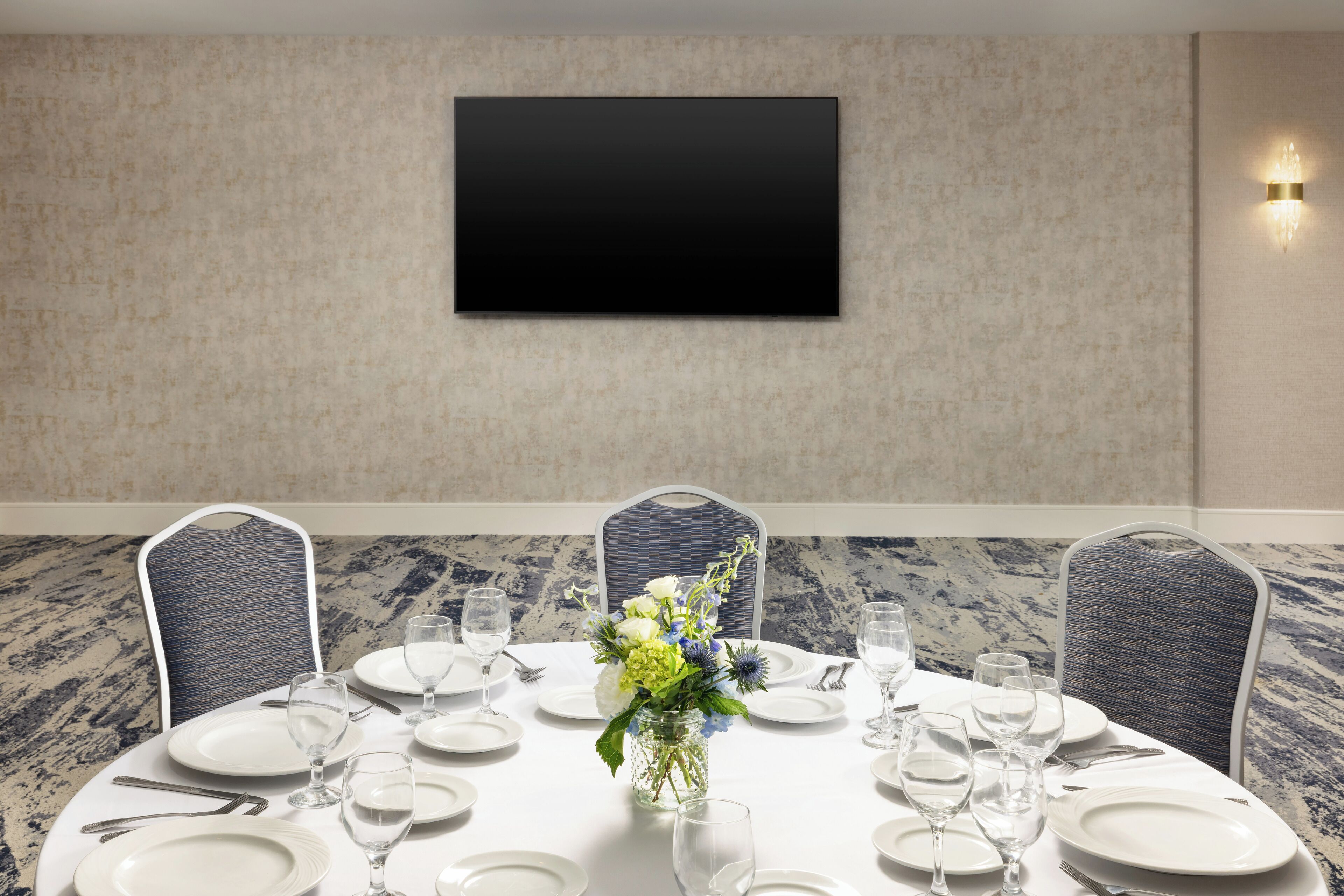Un salón de conferencias con una mesa redonda dispuesta para una reunión, un televisor de pantalla plana montado en la pared y un jarrón decorativo con flores.