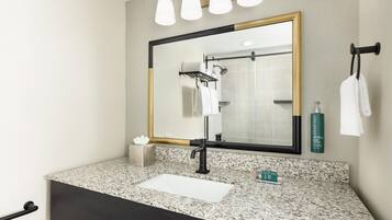 A bathroom with a granite countertop, a black sink, a mirror with a gold frame, a towel rack, and a shower.